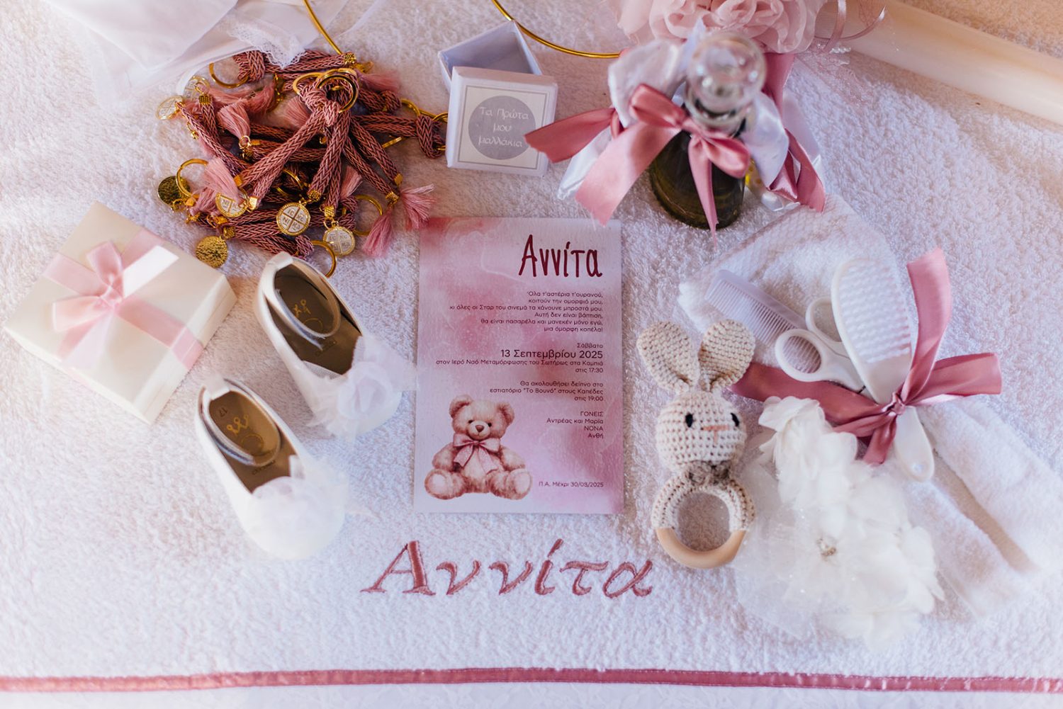 Flat lay of baby girl’s christening items: white shoes, pink invitation, crocheted bunny rattle, and hair accessories on an embroidered towel