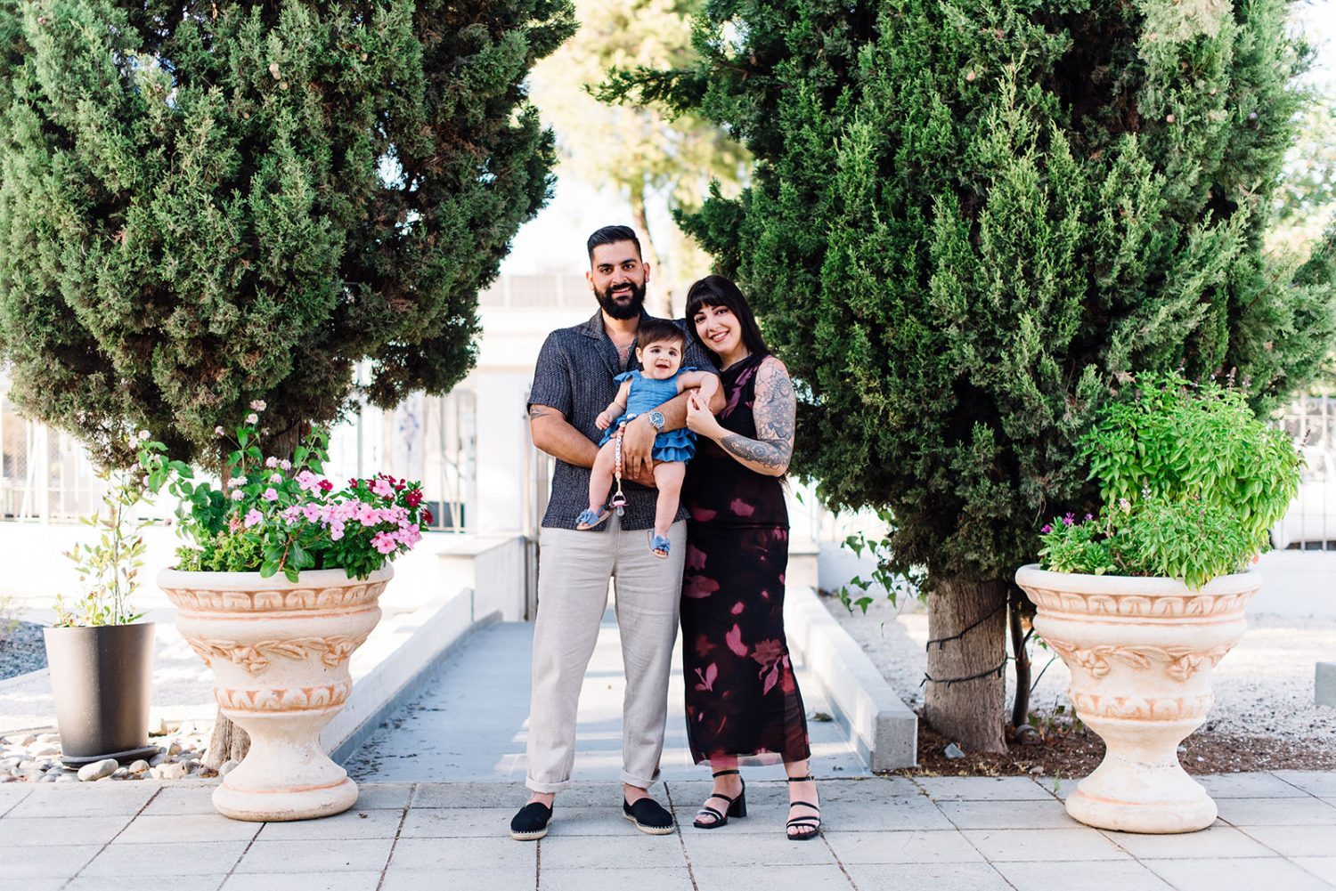 Parents standing outdoors holding their baby girl in a blue dress, between cypress trees and stone planters