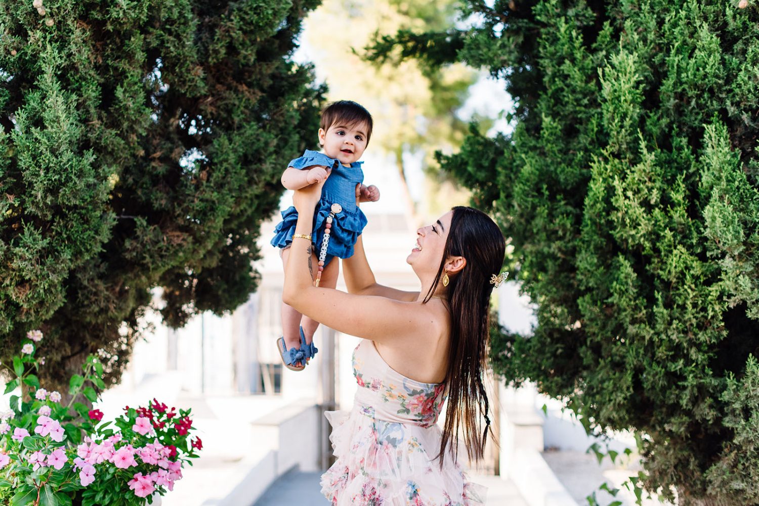 Godmother lifting the baby girl in a blue dress into the air, smiling, with cypress trees behind.