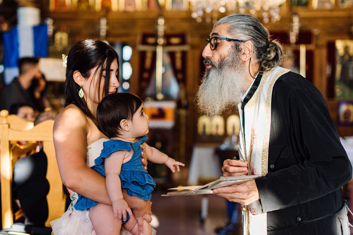 Godmother holding the baby girl as the bearded priest speaks to her during the christening