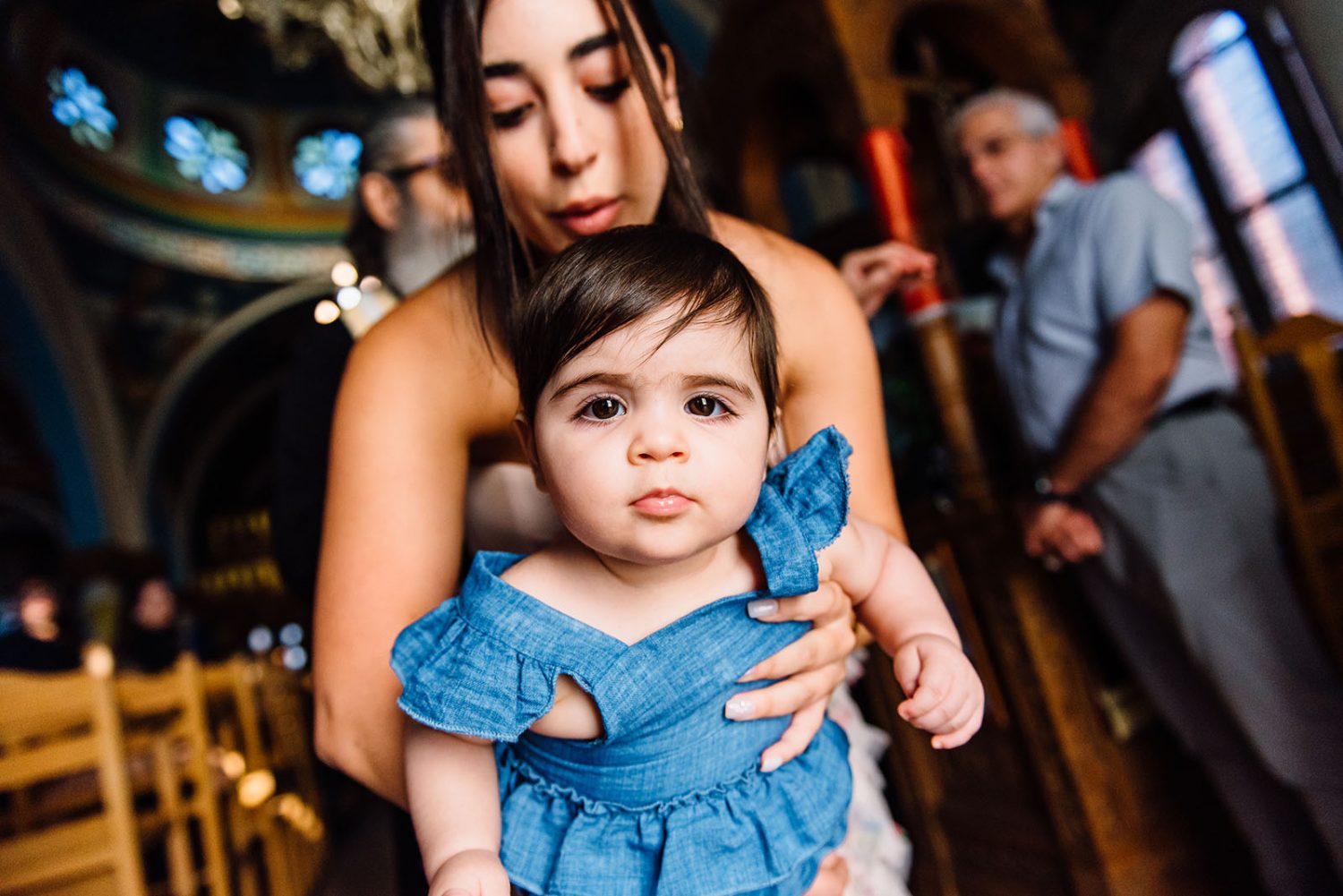 Close-up of the baby girl in a blue dress looking at the camera, held by the godmother inside the church
