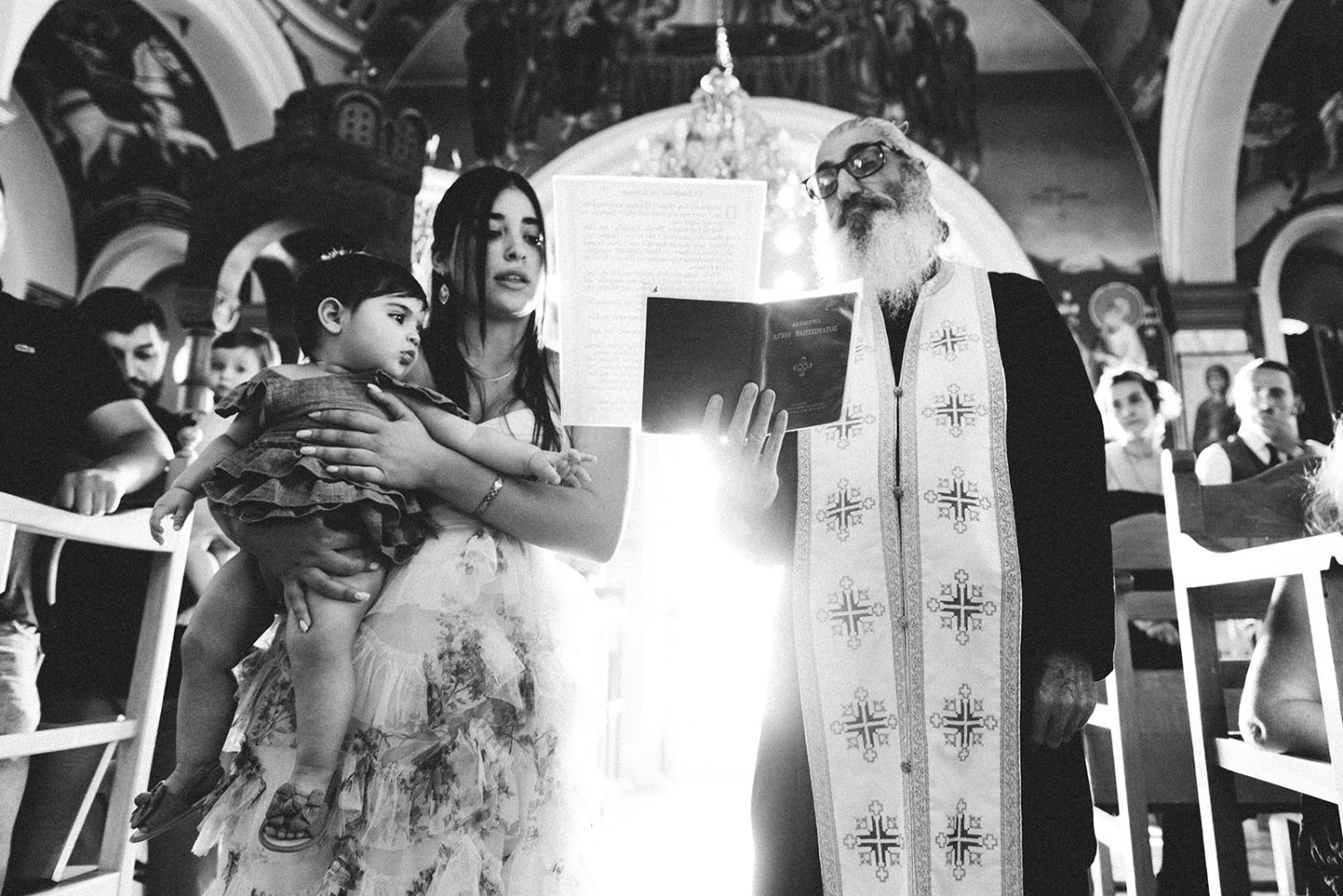 Black and white photo of the godmother holding the baby girl beside the priest as he reads.