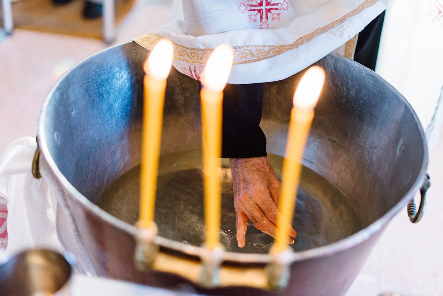 Priest’s hand in the baptism font water, with three lit candles resting on the rim