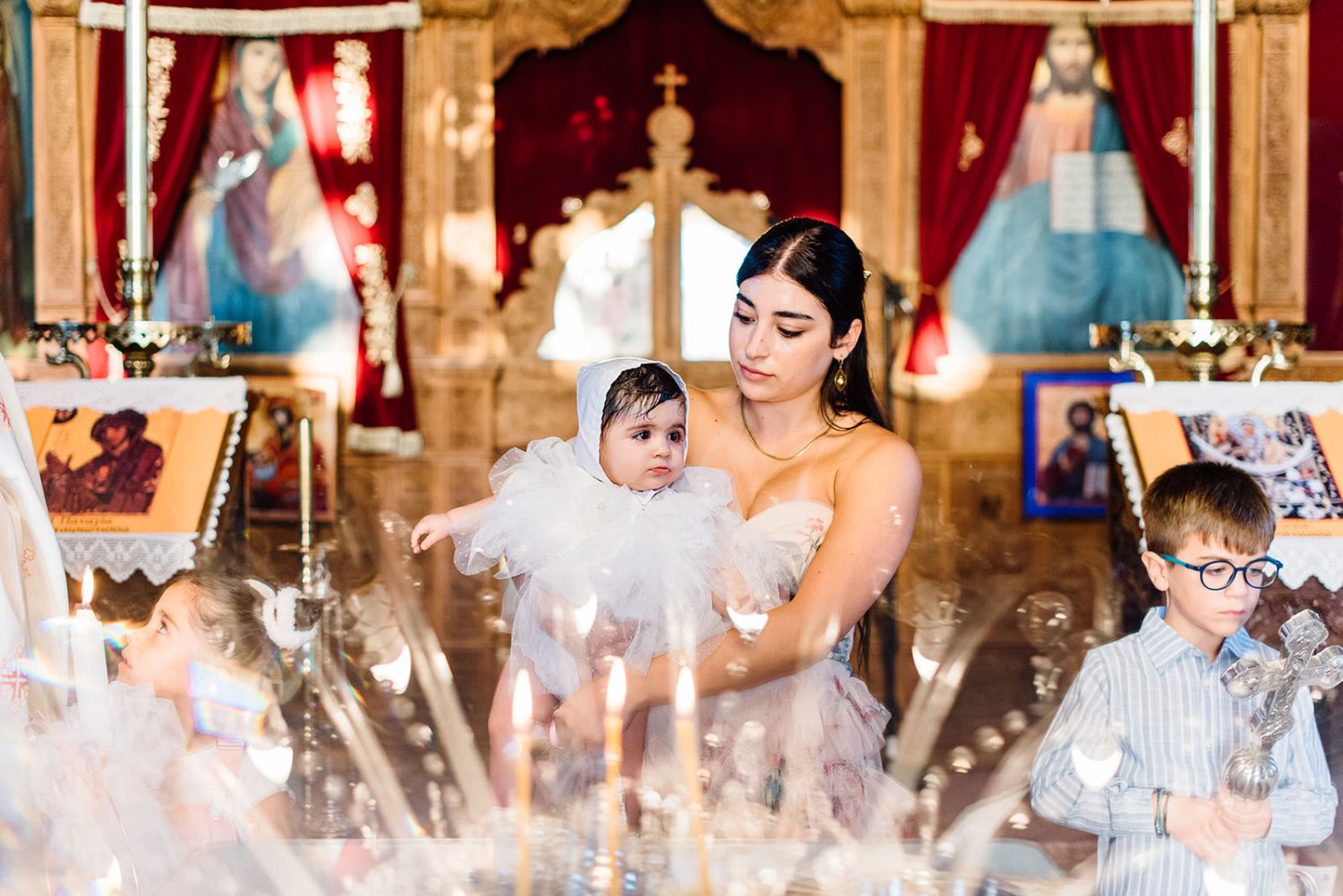 Godmother holding the baby girl in a white dress and bonnet in front of the church iconostasis