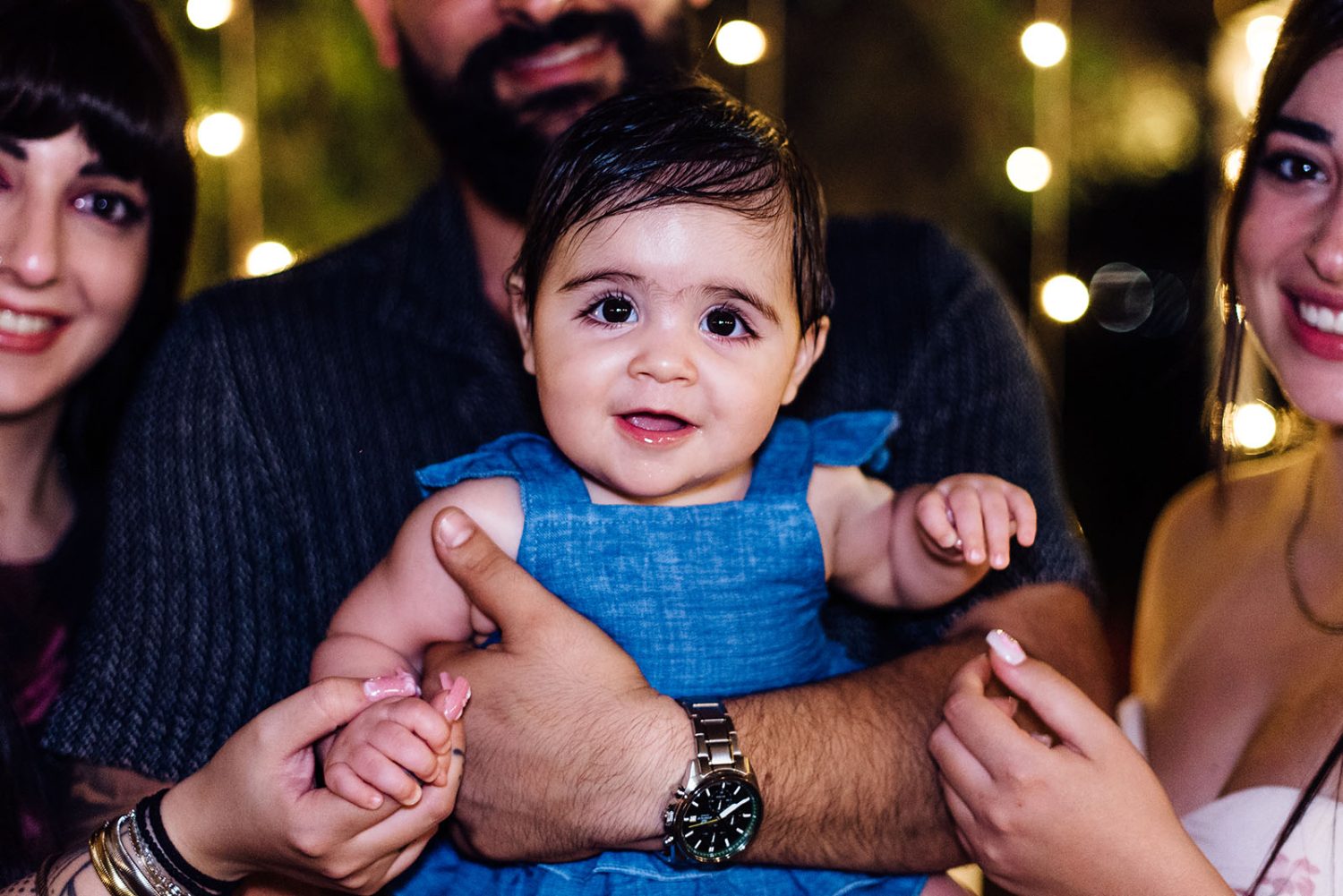 Smiling baby girl in a blue dress held by her father, with family and string lights behind